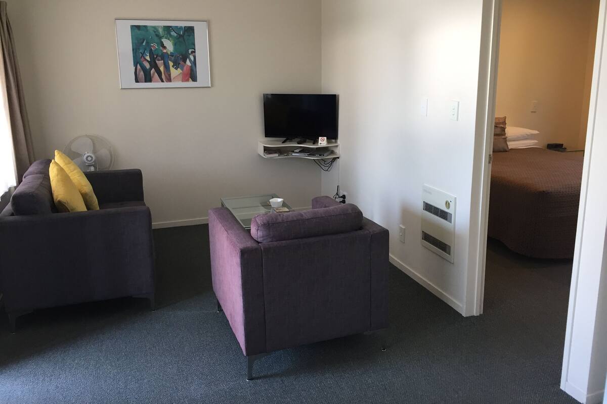A hotel room with a sofa, a small table, a TV, a painting on the wall, and a bed in the adjacent room.