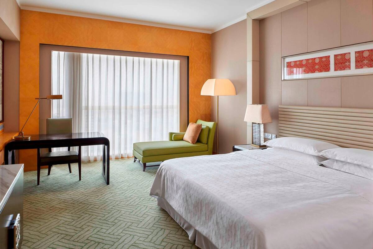 Habitación de hotel con una cama grande, un escritorio, un sillón verde y una ventana con cortinas traslúcidas.