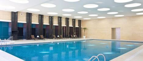 A modern indoor swimming pool with a clear blue water, surrounded by lounge chairs and a ceiling with circular lights.