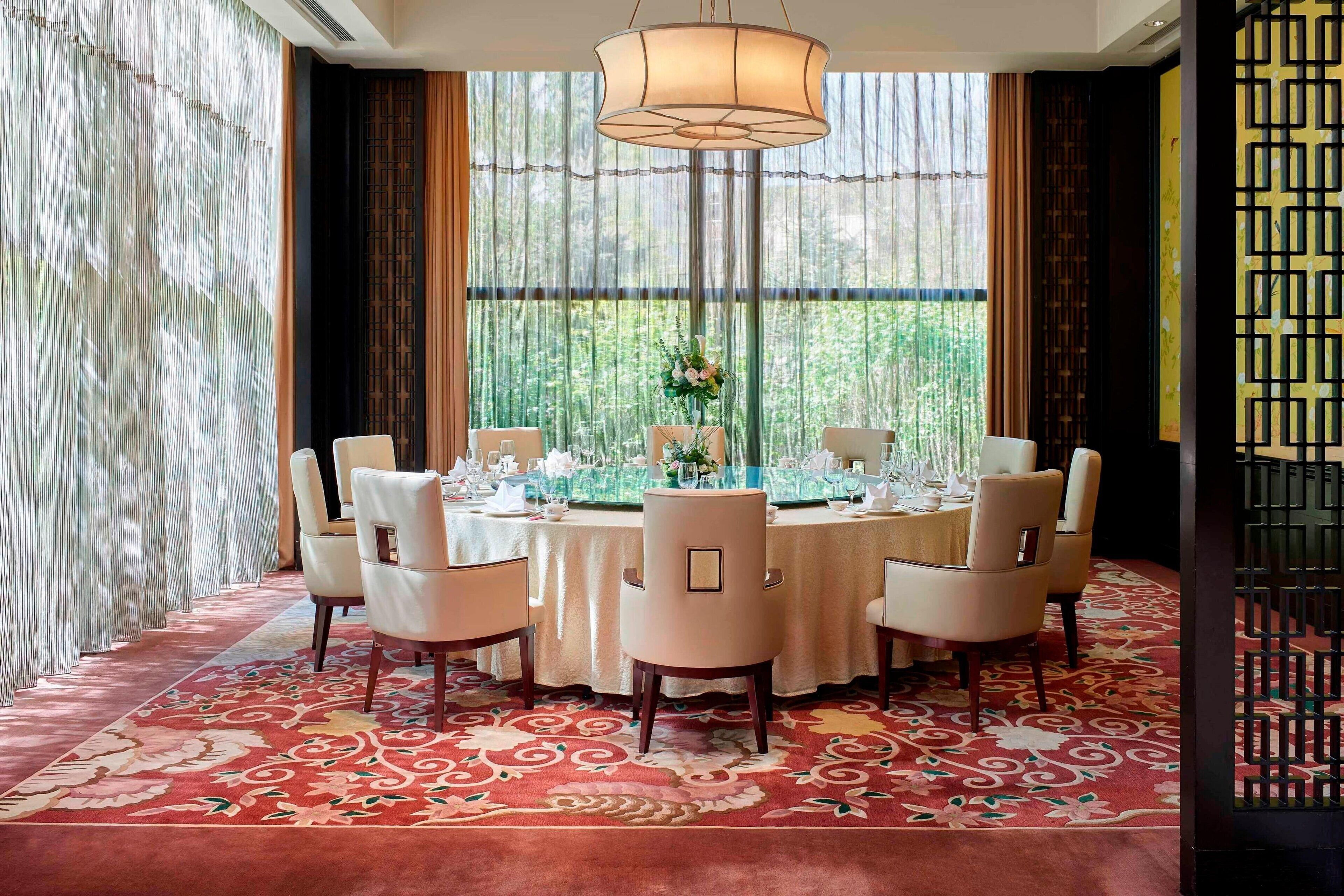 Comedor con una mesa redonda preparada para seis personas, rodeada de sillas, y una ventana grande con cortinas al fondo.