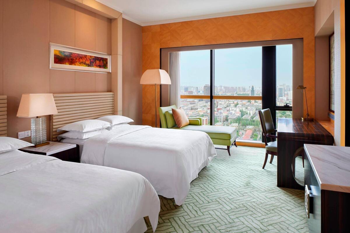Habitación de hotel con dos camas, un escritorio, una silla y un ventanal con vistas a la ciudad.