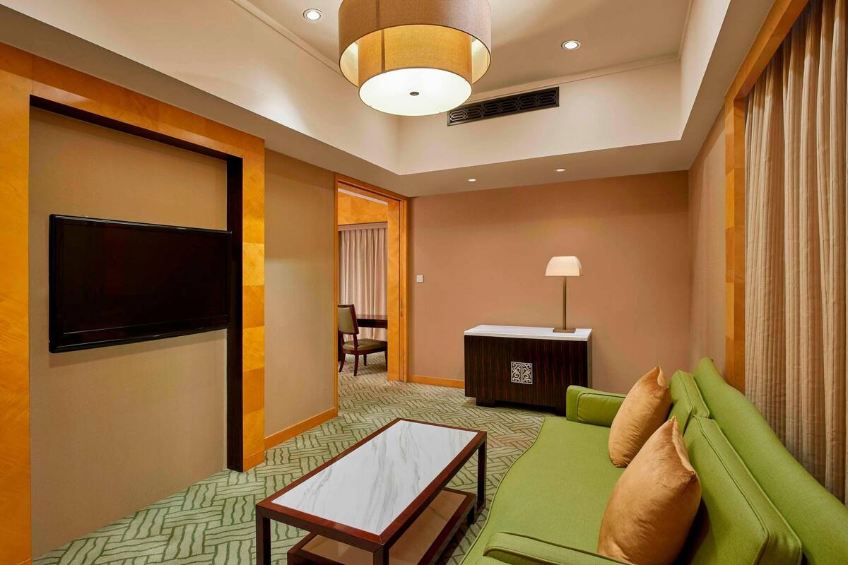 Habitación de hotel con un sofá verde, un televisor de pantalla plana, una mesita y un escritorio con lámpara.