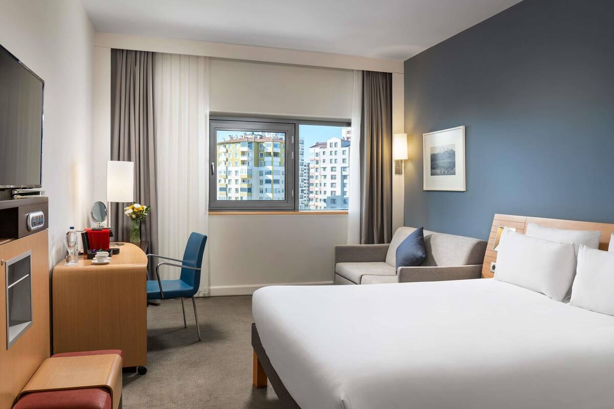A hotel room with a bed, a desk with a lamp, a chair, a TV, and a window with curtains.