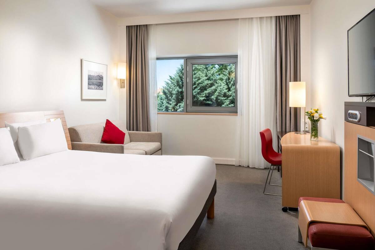A hotel room with a large bed, a desk with a lamp, a chair, and a TV.