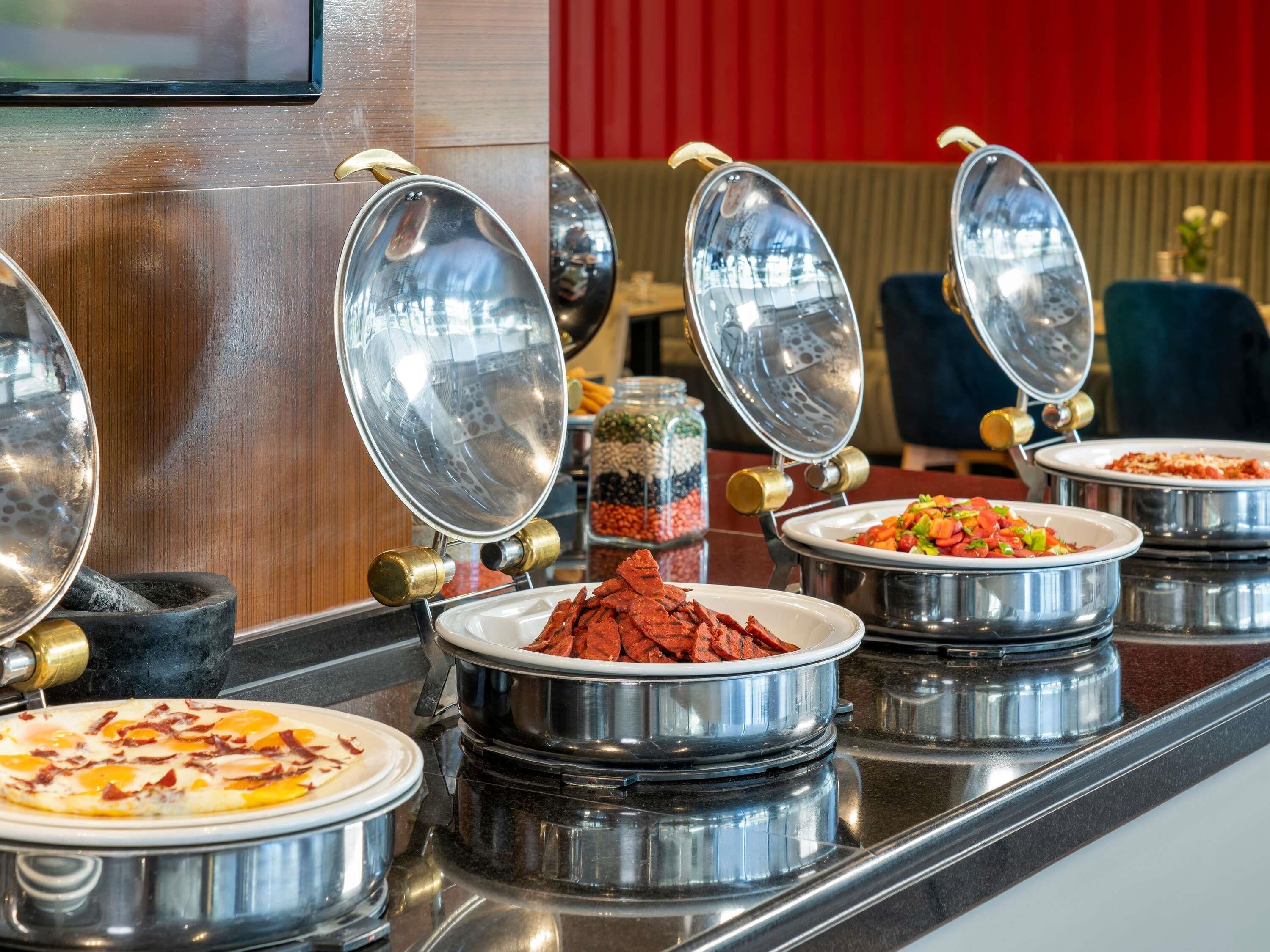 A buffet setup with various food items in metal containers, a coffee machine, and a seating area in the background.
