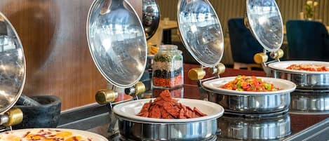 Disposição de buffet com diversos alimentos em recipientes metálicos, uma máquina de café e uma área de estar ao fundo.
