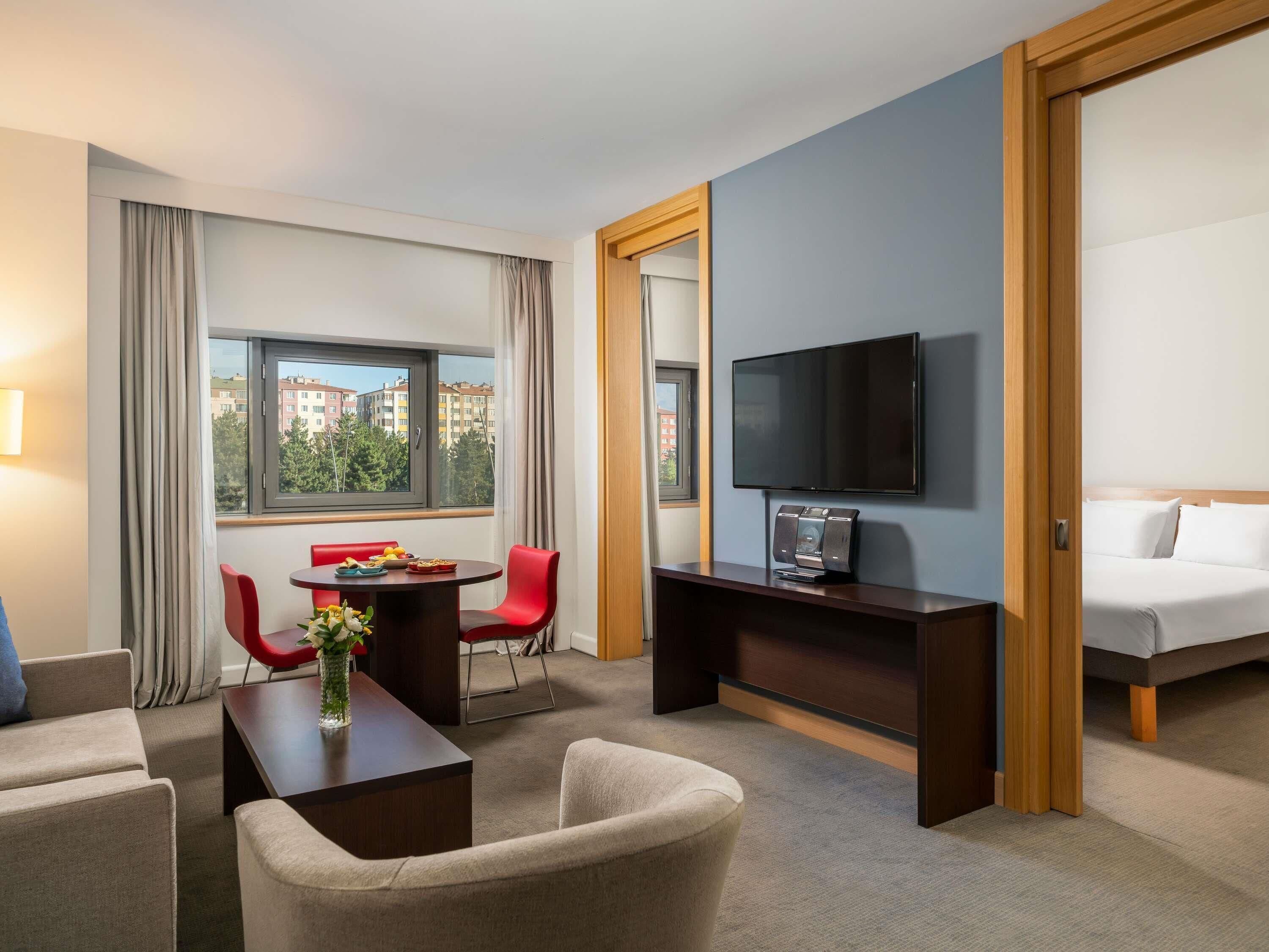 A hotel room with a bed, a TV, a desk with a chair, a small table with a vase, and a window with curtains.