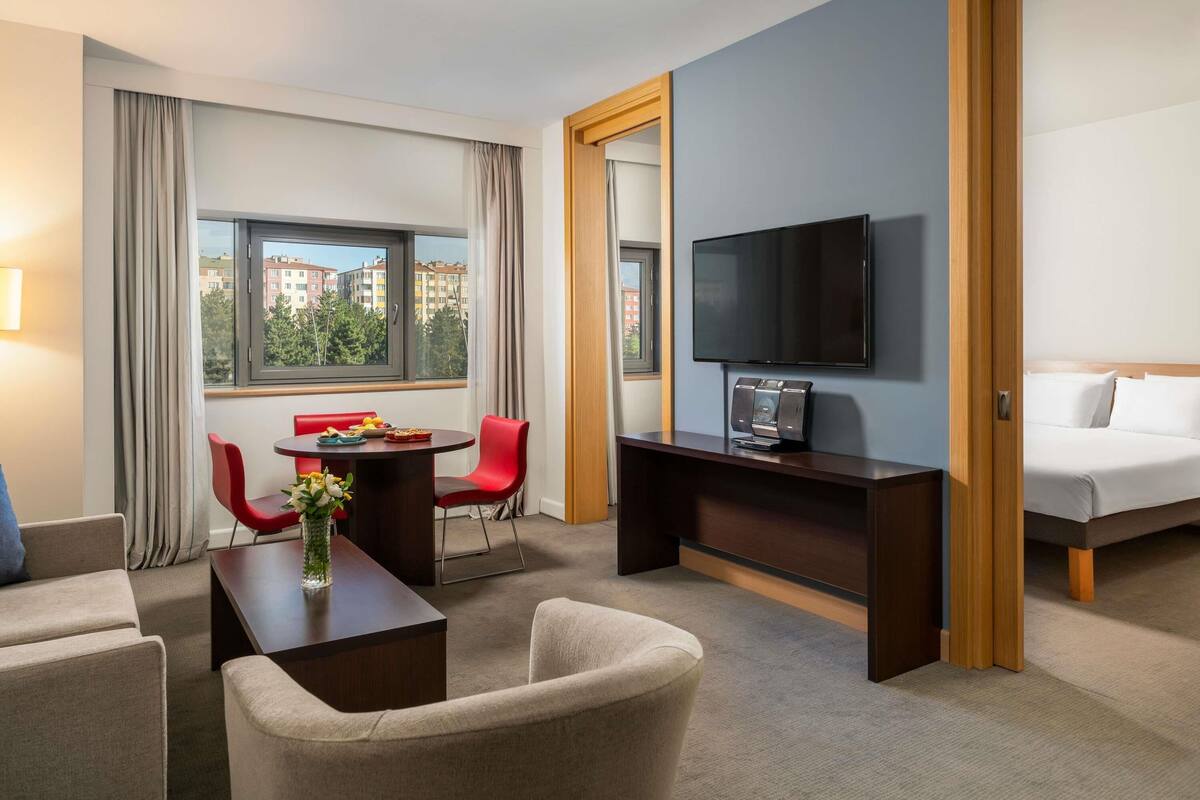 A hotel room with a bed, a TV, a desk with a chair, a small table with a vase, and a window with curtains.