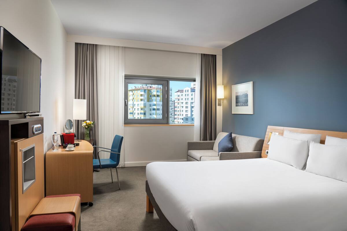 A hotel room with a bed, a desk with a TV, a chair, a sofa, and a window with a city view.