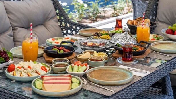 Tafel gedekt met diverse gerechten, waaronder salades, vleeswaren en drankjes, in een terras met rieten meubels en uitzicht op een zwembad en groen.