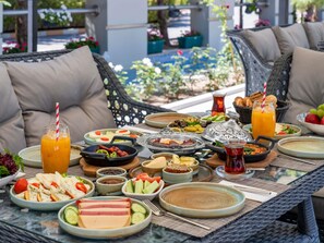 Ein Tisch ist reichhaltig mit Speisen und Getränken gedeckt, darunter Salate, Fleisch und Getränke. Das Esszimmer befindet sich im Freien, mit Korbmöbeln und Blick auf Pool und Grünflächen.