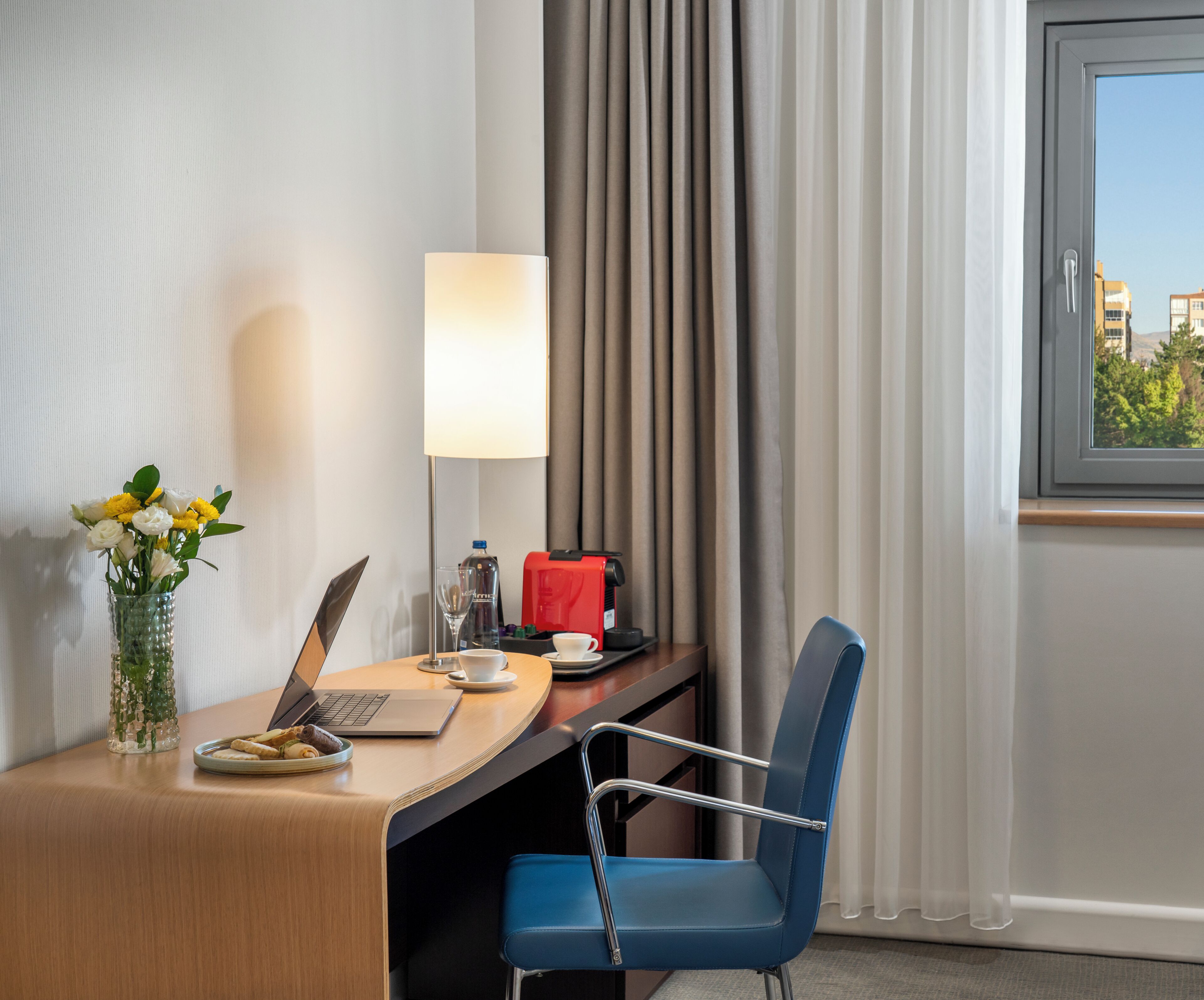 A hotel room with a desk, laptop, coffee cup, tea cup, and a vase with flowers.