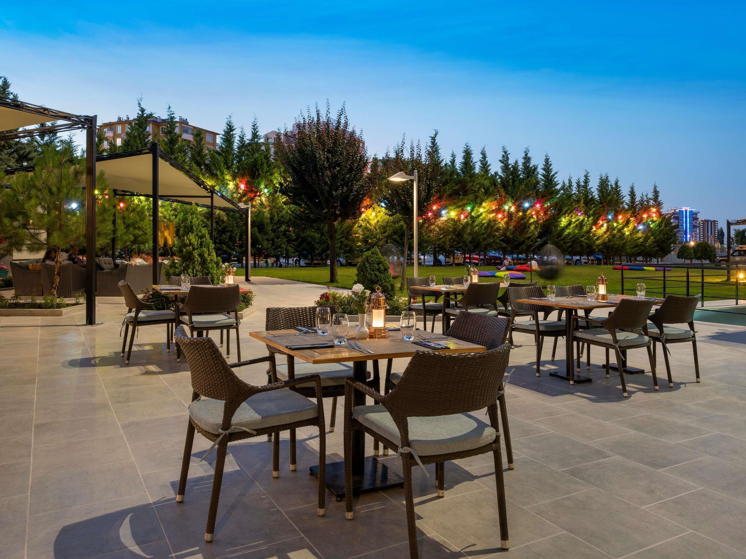 Un espacio de comedor al aire libre con mesas y sillas, rodeado de árboles y vistas a edificios.