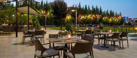 Un espacio de comedor al aire libre con mesas y sillas, rodeado de árboles y vistas a edificios.