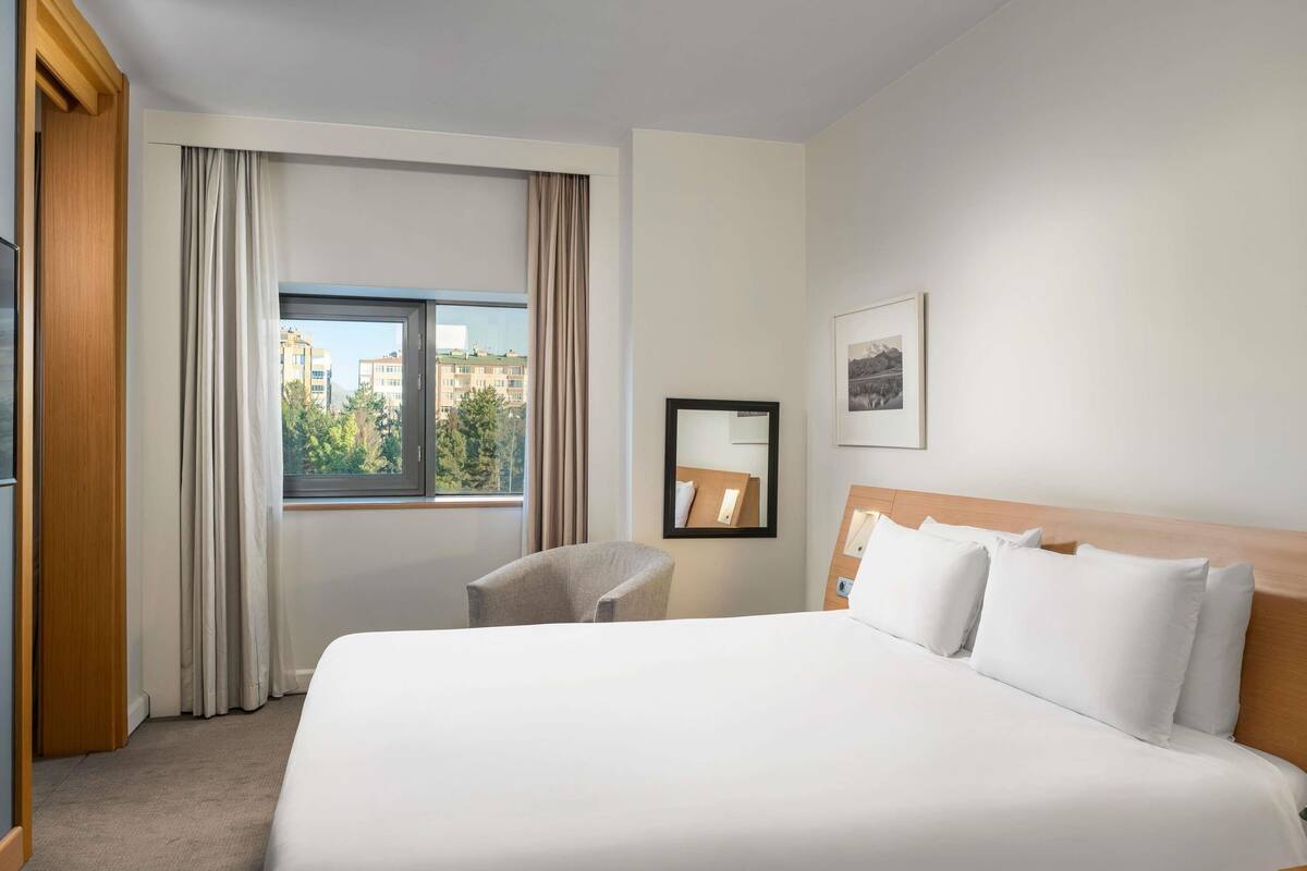 A hotel room with a large bed, a chair, a window with curtains, a framed mirror, and a picture on the wall.