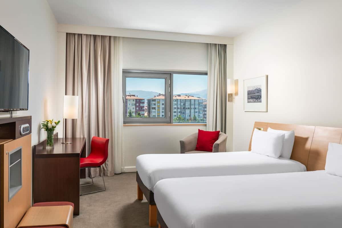 A hotel room with two beds, a desk with a TV, a chair, a window with a view of buildings, and a lamp.
