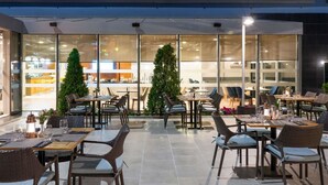 Een terras met meerdere tafels en stoelen, grote ramen en uitzicht op het interieur.
