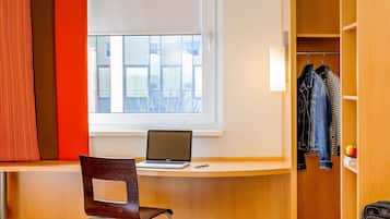 A hotel room with a bed, a desk with a laptop, a chair, a wardrobe, and a window with a view of buildings.
