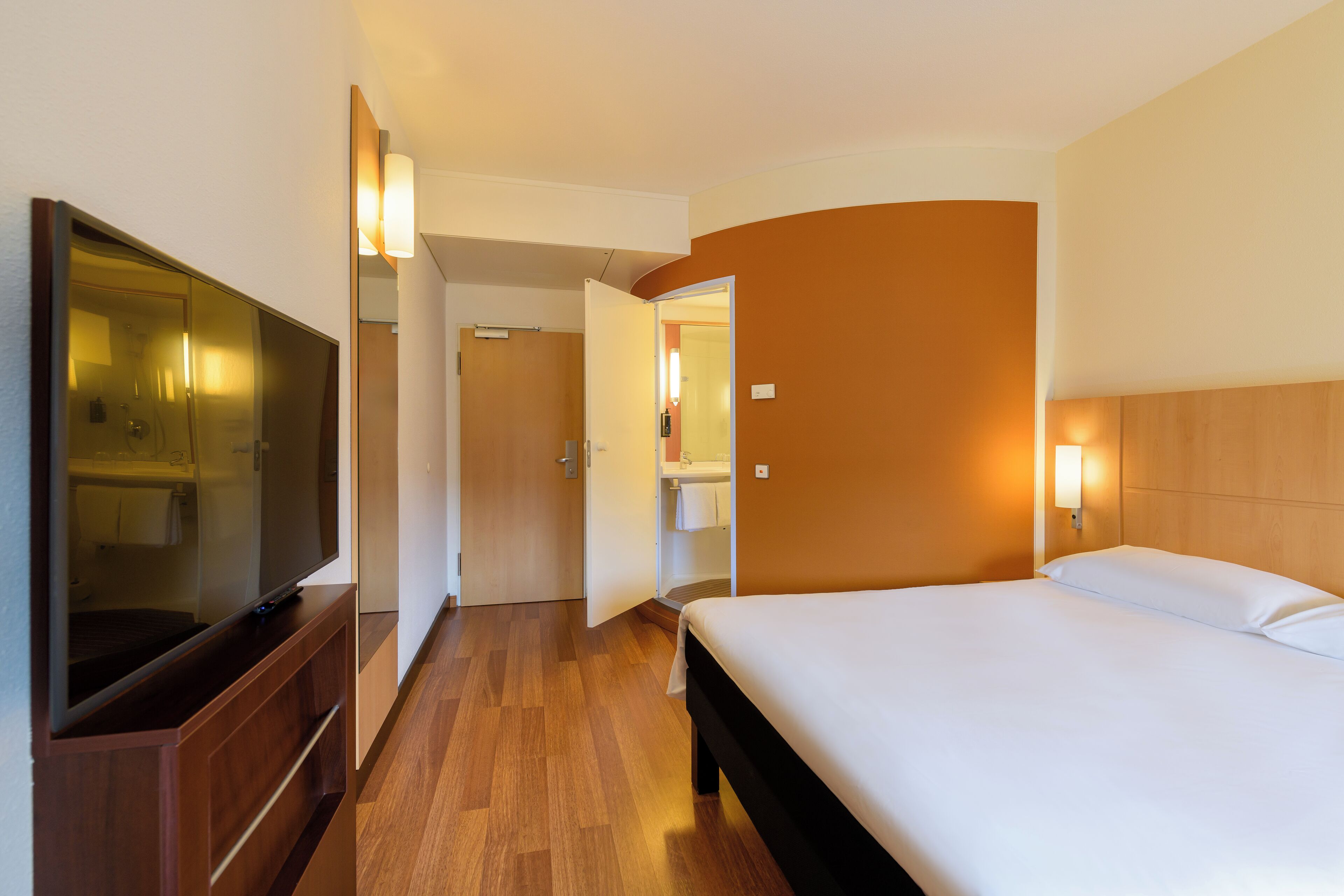 Habitación de hotel con cama, televisión, espejo y baño visible a través de una puerta abierta.