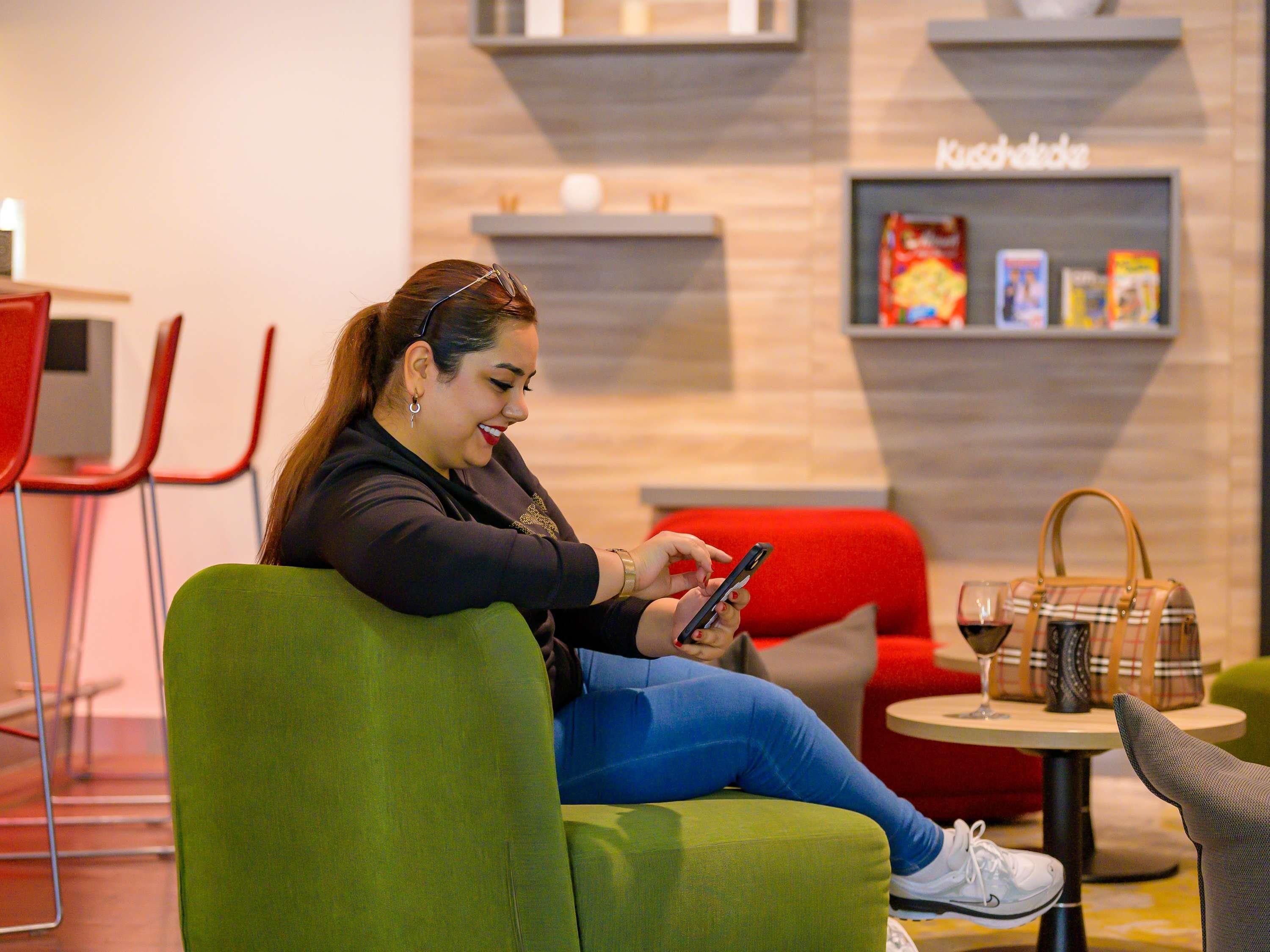A person seated on a green chair, using a smartphone. There are red chairs and a small round table with a handbag and a wine glass on it.