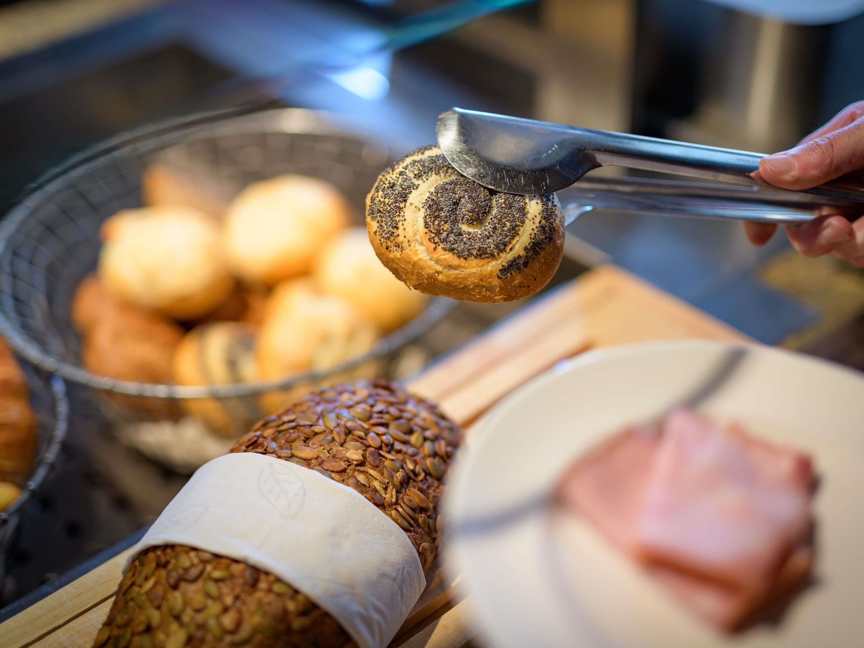 A hand holding tongs over a poppy seed bun, with a basket of buns and a loaf of seeded bread in the background.