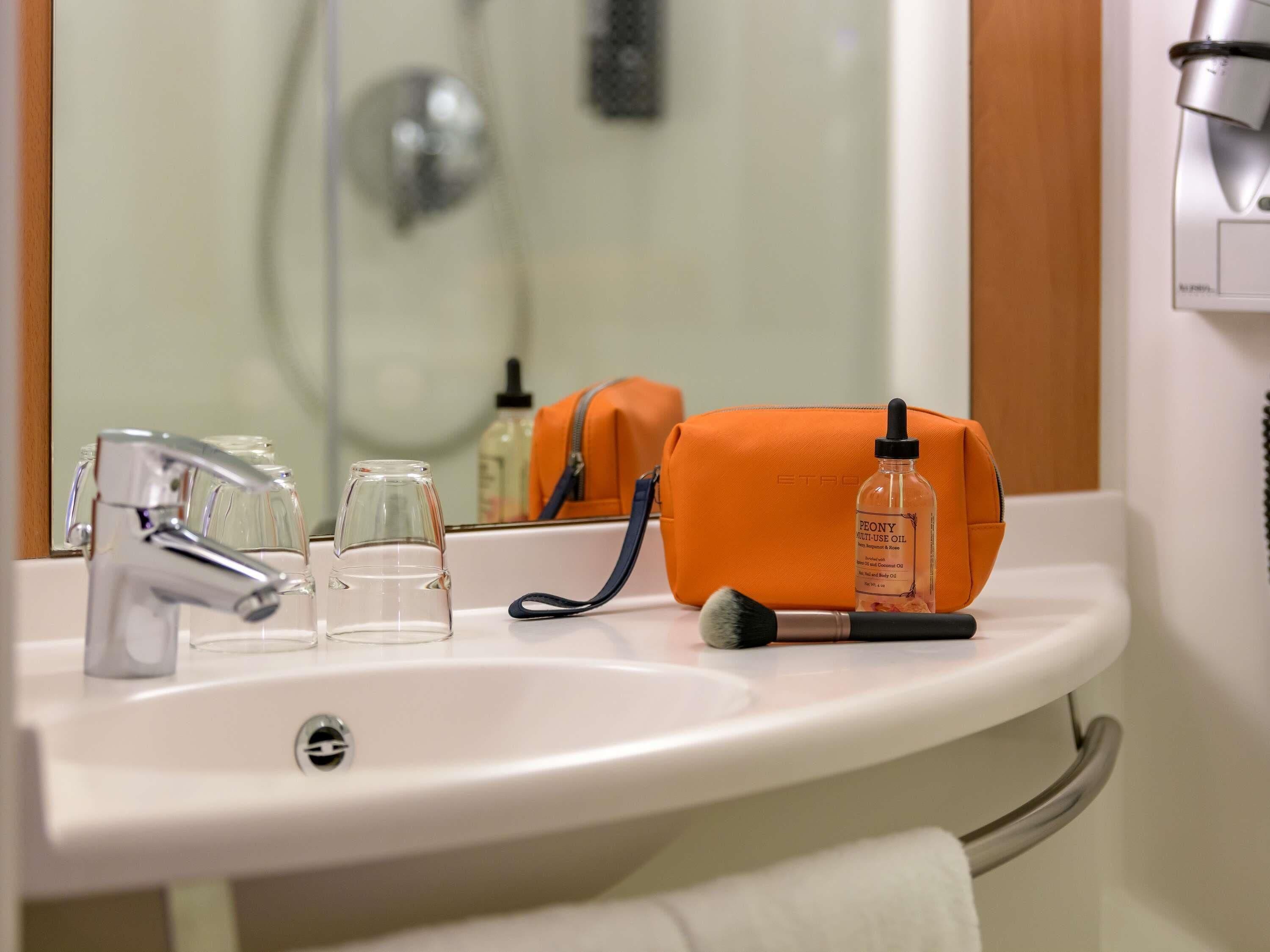 Un lavabo de salle de bain avec un miroir, un robinet, un verre, une bouteille d’huile et un sac orange.