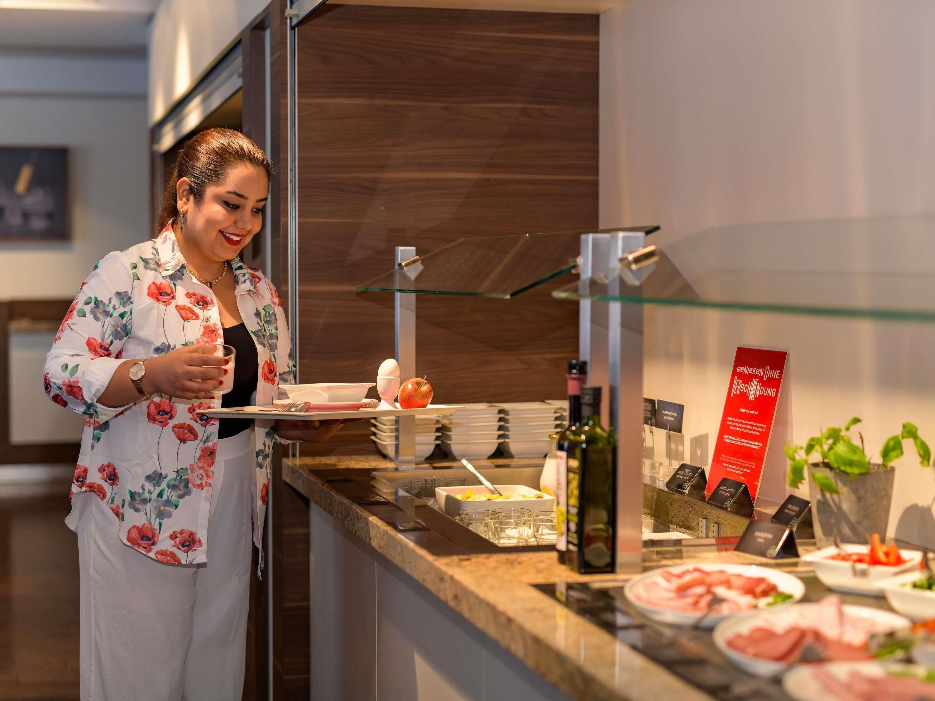 A person serving food at a counter with a glass display case, a menu stand, and various food items.