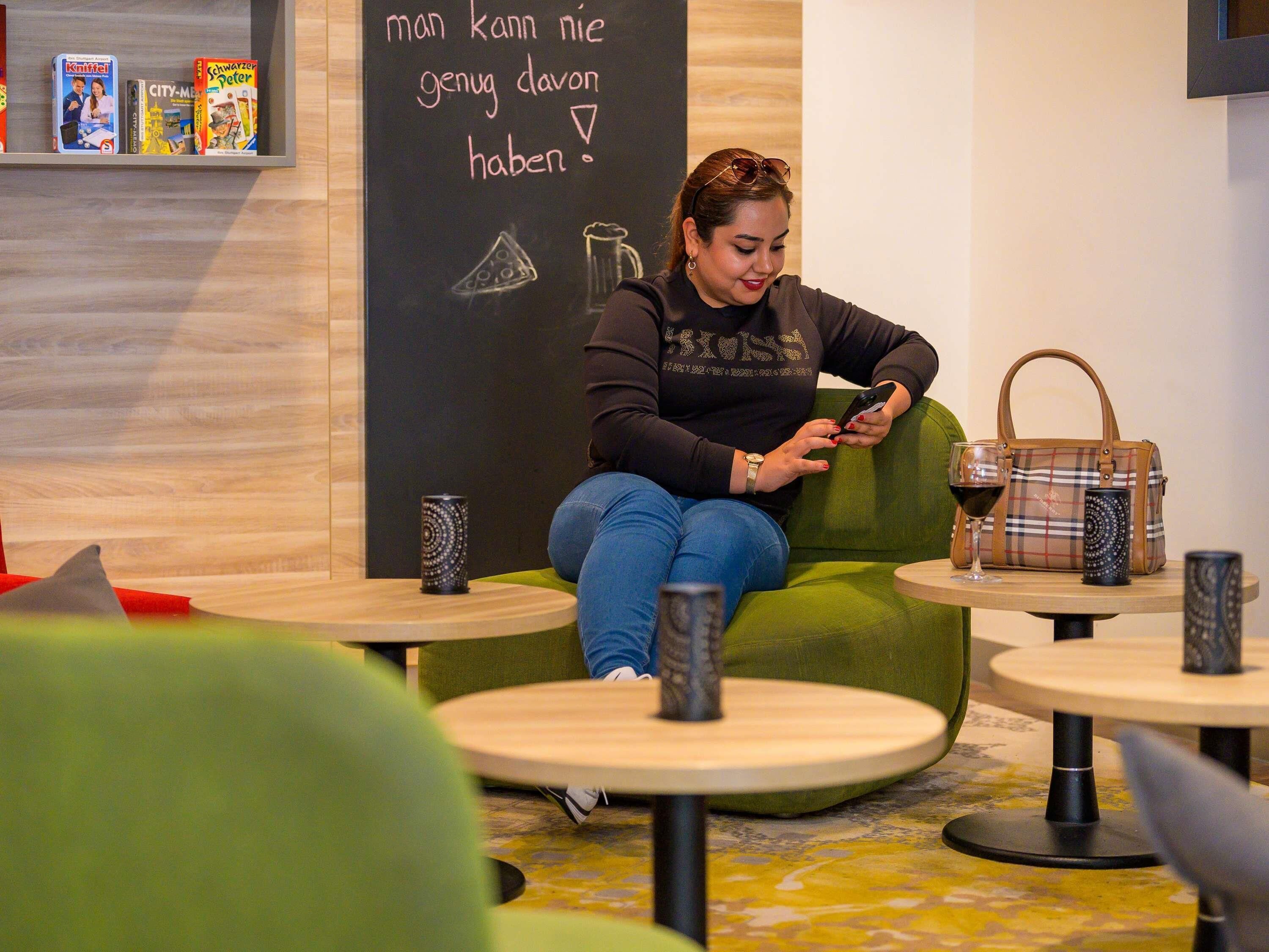 A person seated on a green couch in a modern interior with a chalkboard wall, a small table with a glass and a bottle, and a shelf with books.
