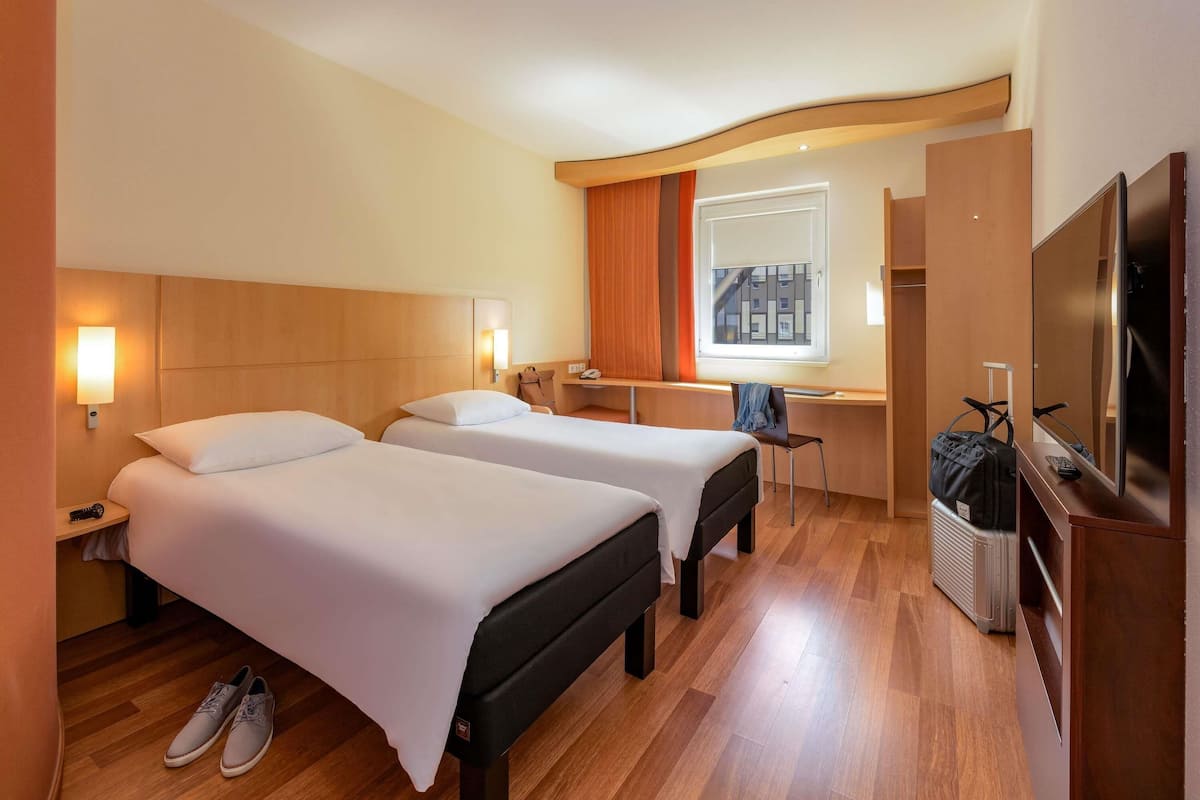 Une chambre d’hôtel avec deux lits, un bureau, une chaise, une télévision et une fenêtre donnant sur des immeubles.