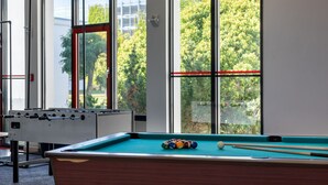 Une table de billard avec des billes et des queues, une table de baby-foot, et de grandes fenêtres donnant sur un paysage verdoyant.
