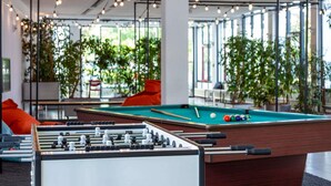 Une table de billard et une table de baby-foot dans une pièce dotée de grandes fenêtres et agrémentée de plantes d’intérieur.
