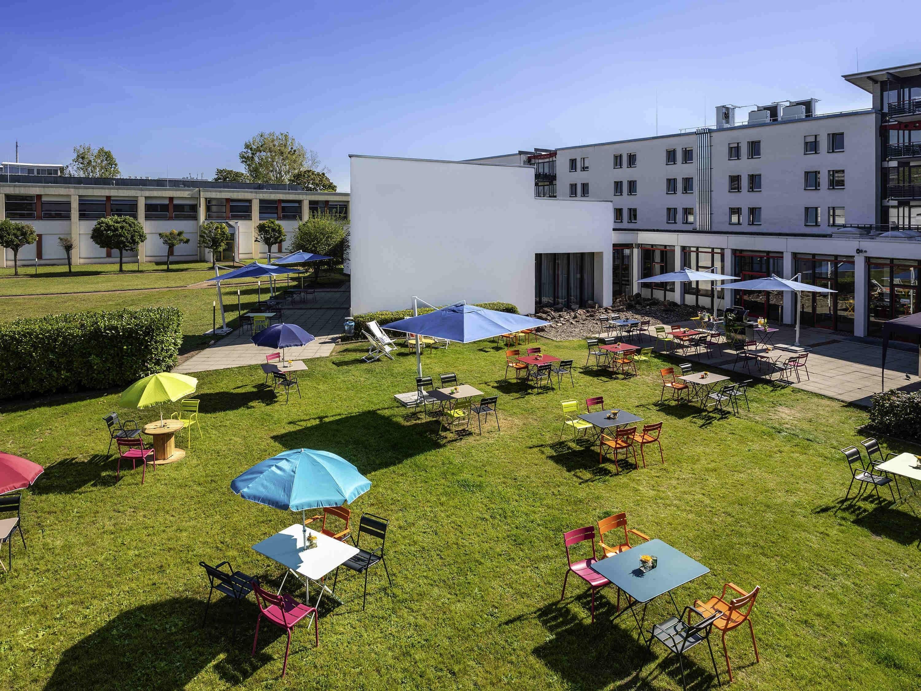 Een buitenterras met meerdere tafels en stoelen, parasols en een gebouw op de achtergrond.