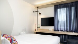 Une chambre d’hôtel moderne équipée d’un lit, d’un téléviseur à écran plat fixé au mur, d’un bureau avec une lampe et d’une fenêtre avec des rideaux.