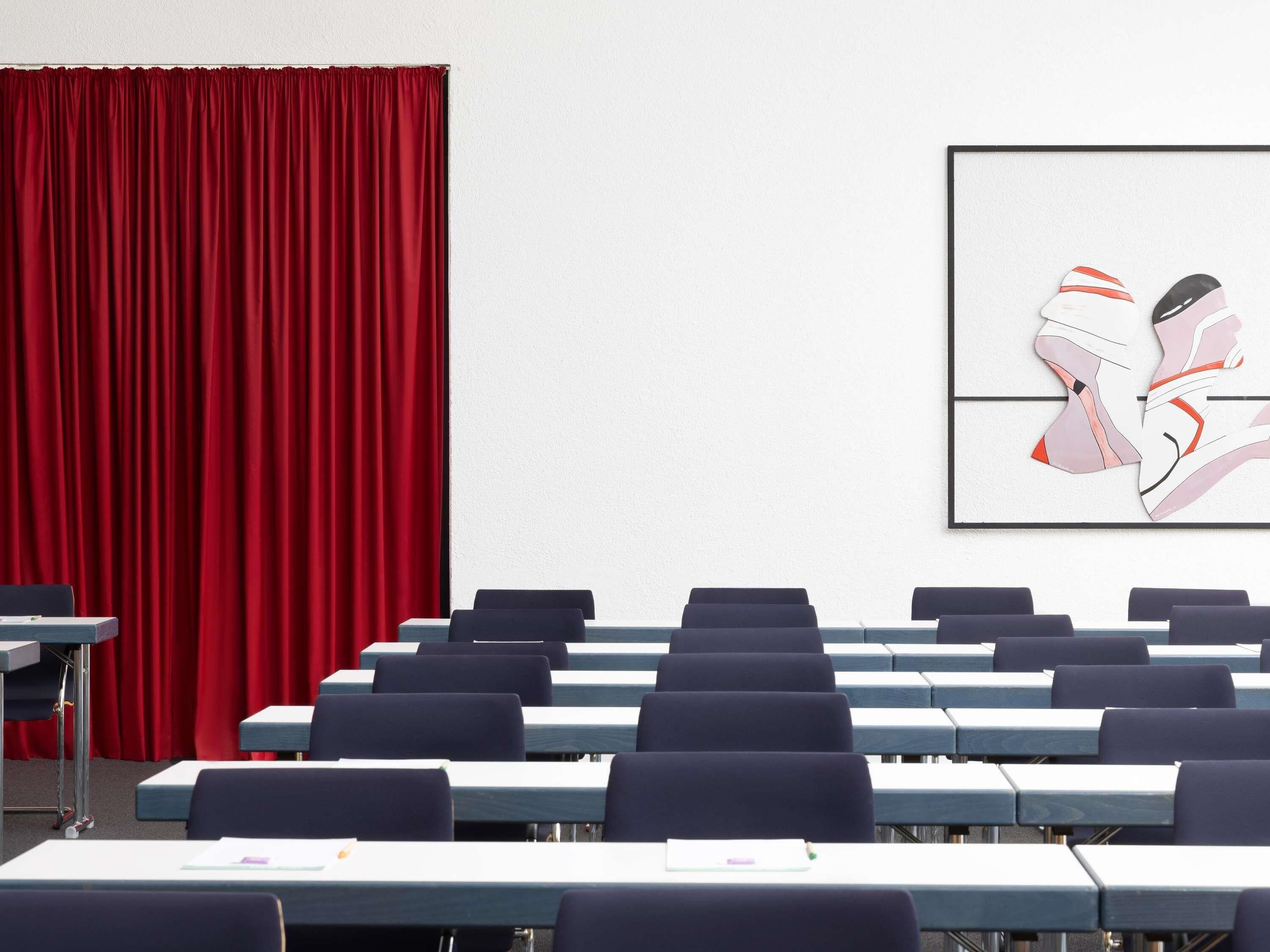 A conference room with rows of tables and chairs, a red curtain, and a framed abstract art piece on the wall.