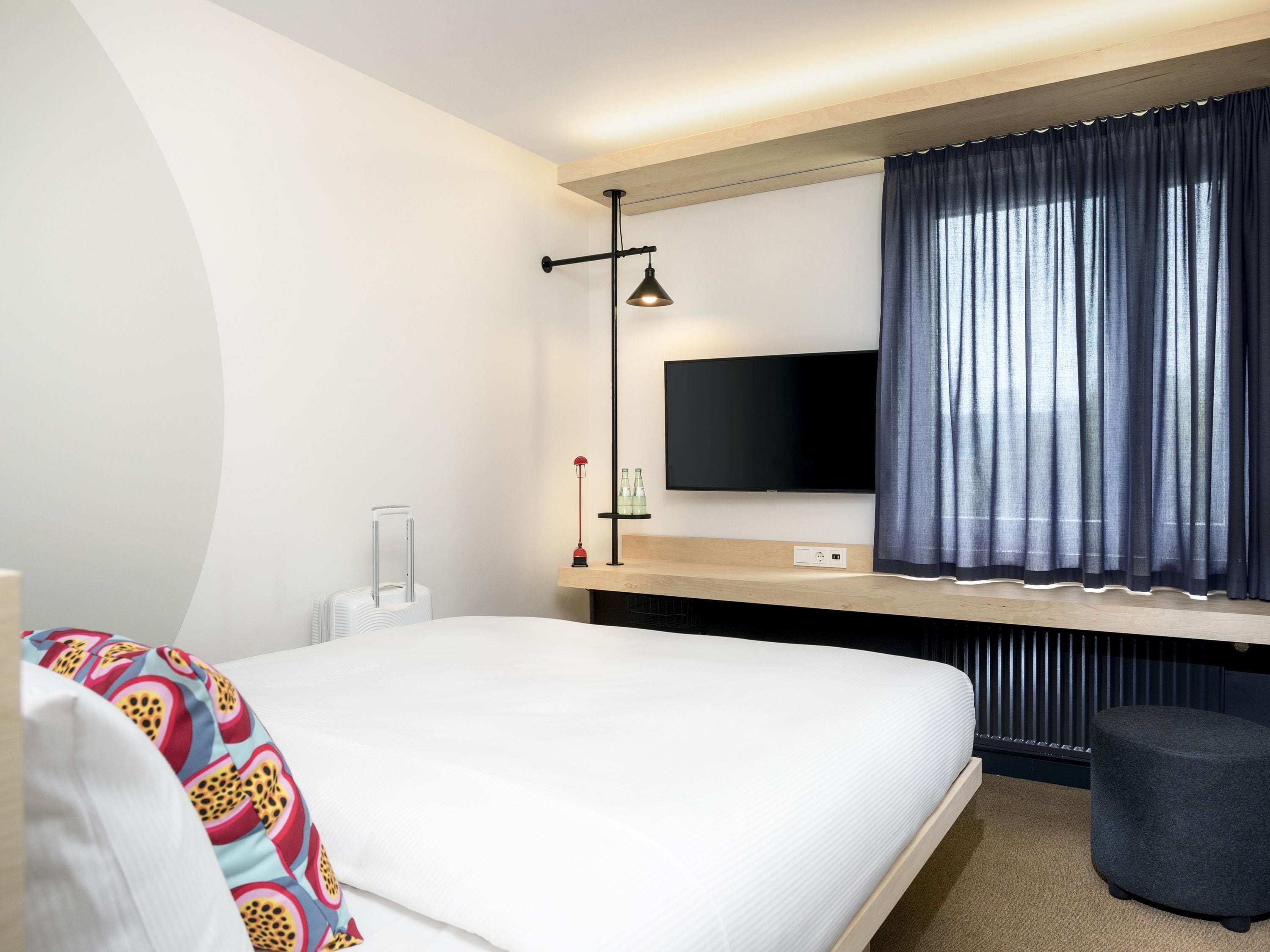 Habitació d'hotel moderna amb llit, televisió de pantalla plana penjada a la paret, escriptori amb làmpada i finestra amb cortines.