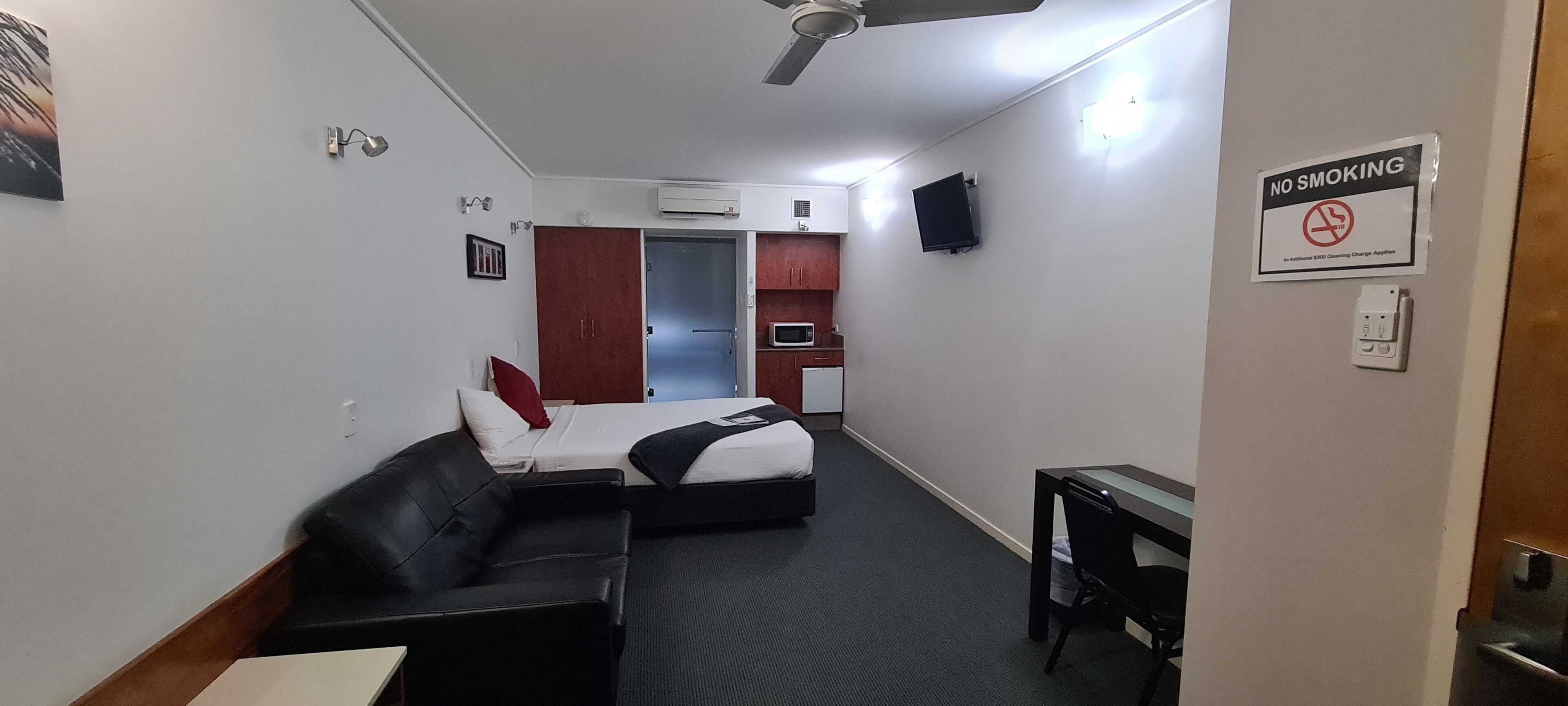 A compact hotel room with a bed, a sofa, a desk, a microwave, and a kitchenette. A 'No Smoking' sign is visible on the wall.