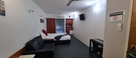 A compact hotel room with a bed, a sofa, a desk, a microwave, and a kitchenette. A 'No Smoking' sign is visible on the wall.