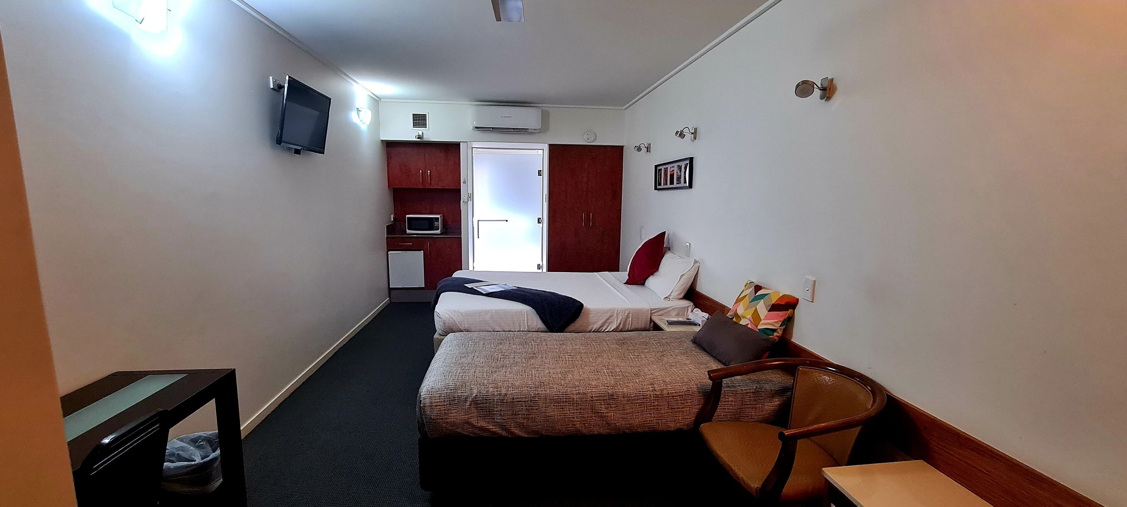 A compact hotel room with two beds, a small kitchenette, a TV, and a single chair.