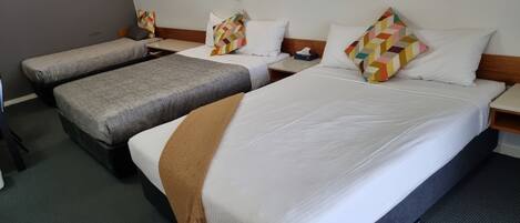 Een hotelkamer met drie eenpersoonsbedden, elk met een ander kussendesign. Tussen de bedden staat een klein tafeltje, en een nachtkastje met erin een zakdoos.