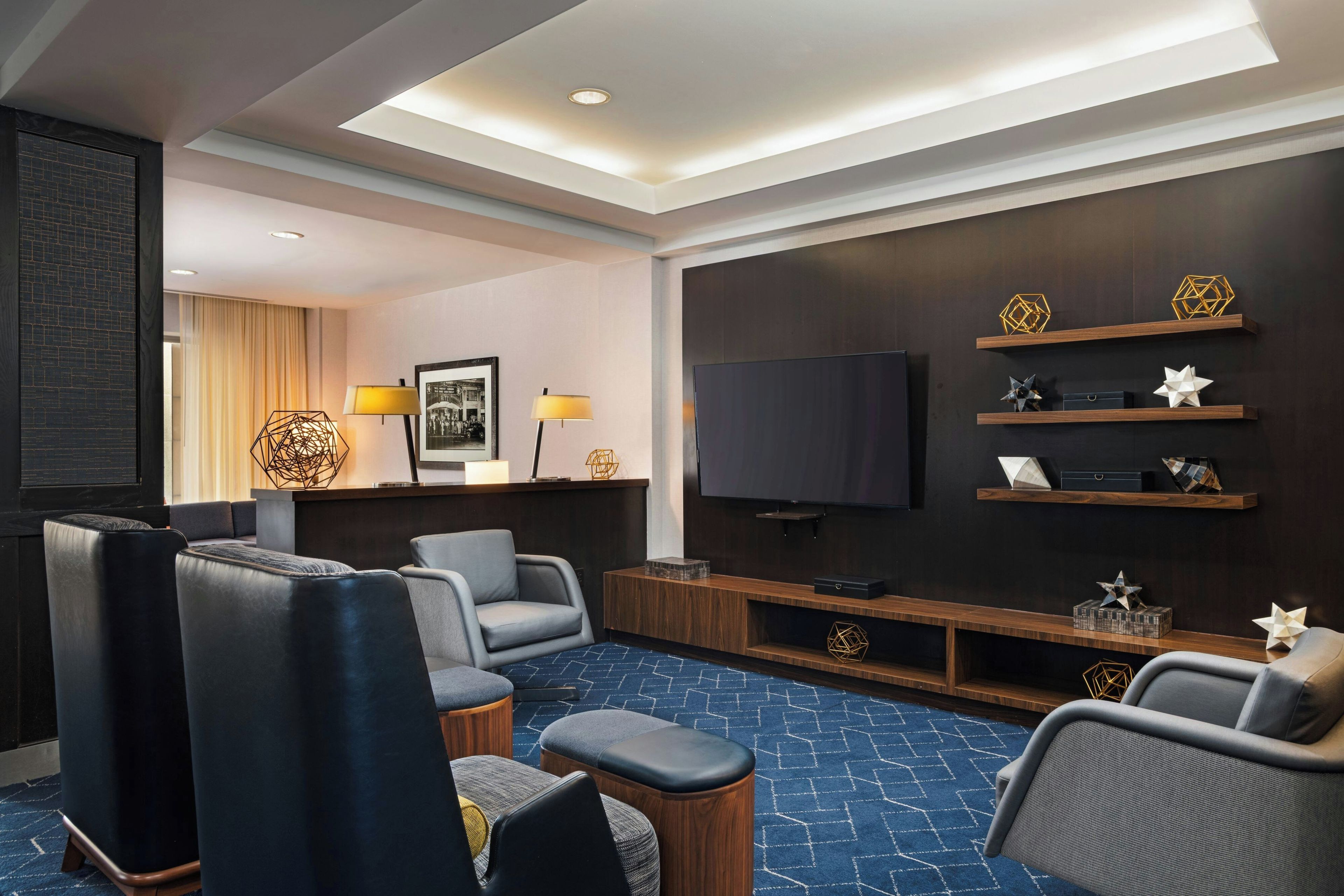 A modern hotel room with a flat-screen TV, leather seating, a patterned carpet, and decorative wall shelves.