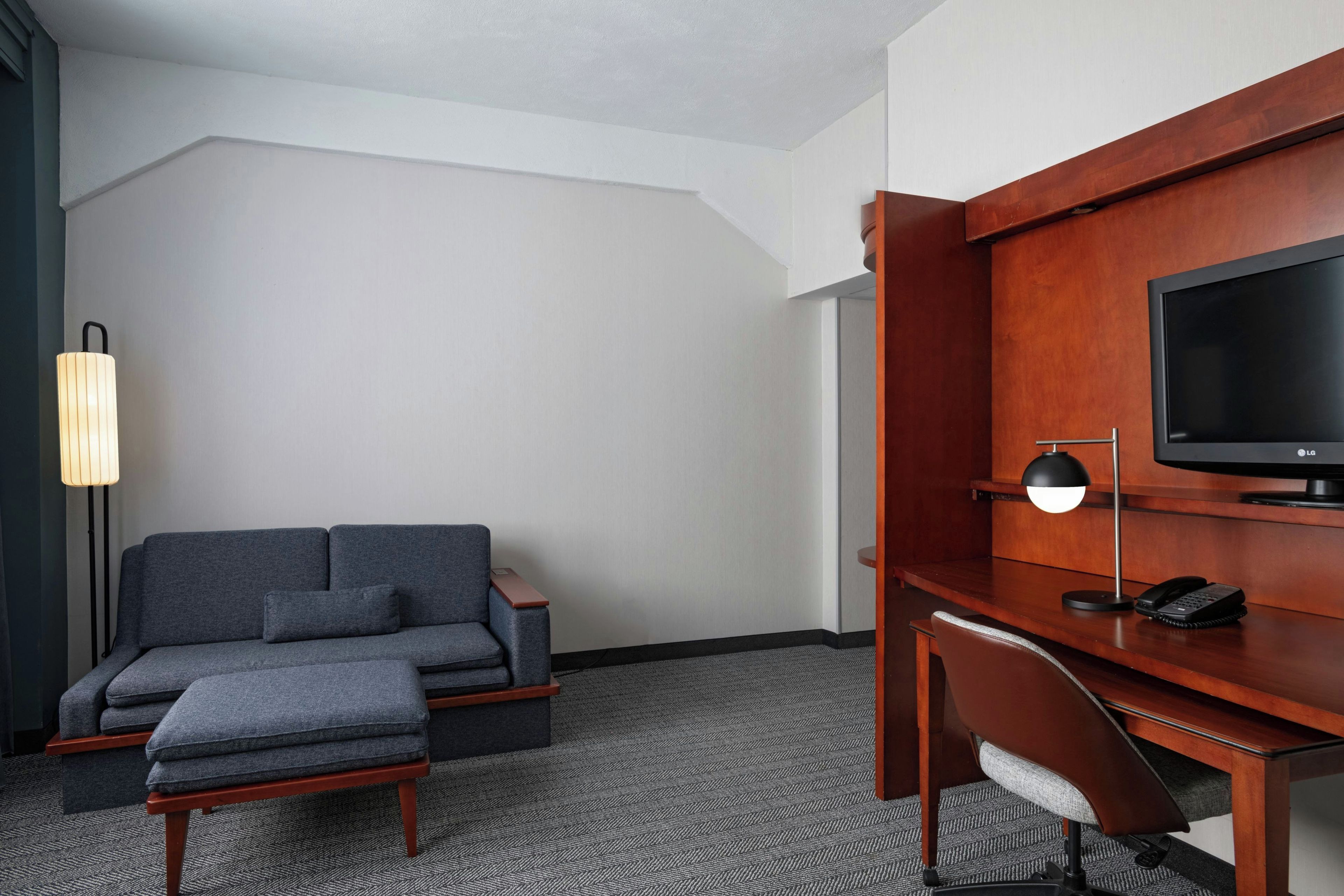 Habitación de hotel con un sofá gris, un escritorio de madera, una televisión y una lámpara.