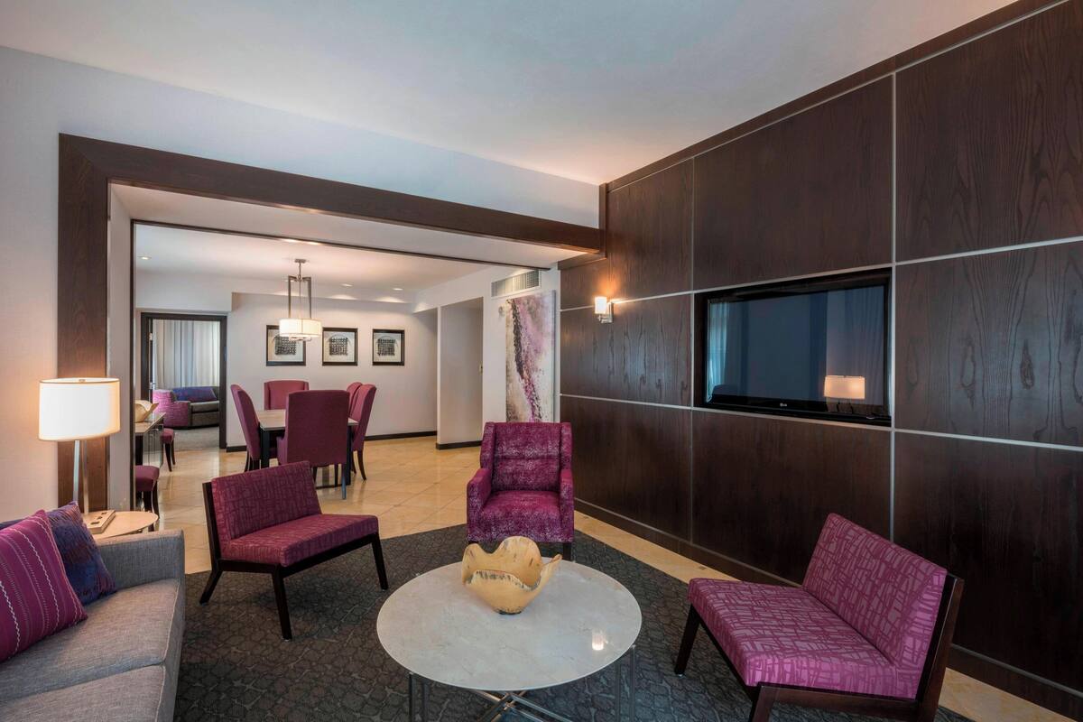 A hotel lobby with a round table, purple armchairs, a sofa, a TV mounted on the wall, and a dining area in the background.