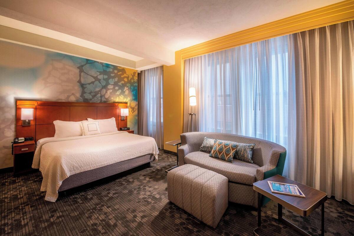 A hotel room with a large bed, a sofa, a small table, and a window with curtains.