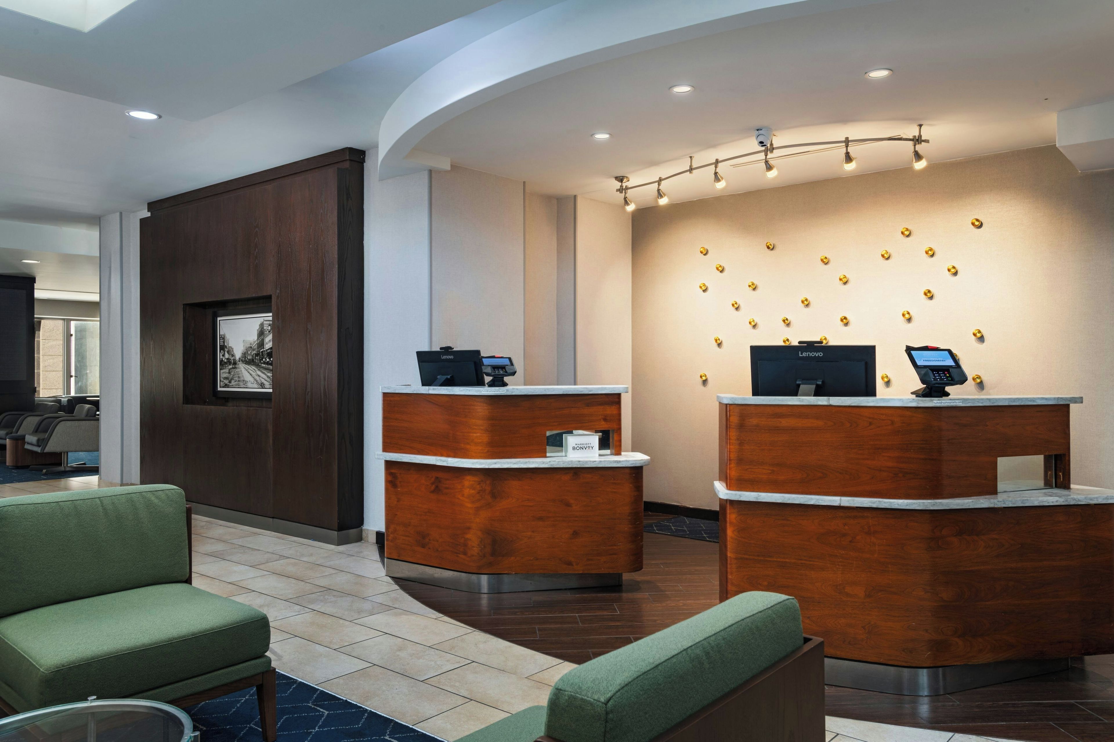 A hotel reception area with two reception desks, a framed picture on the wall, and a seating area with green upholstered chairs.