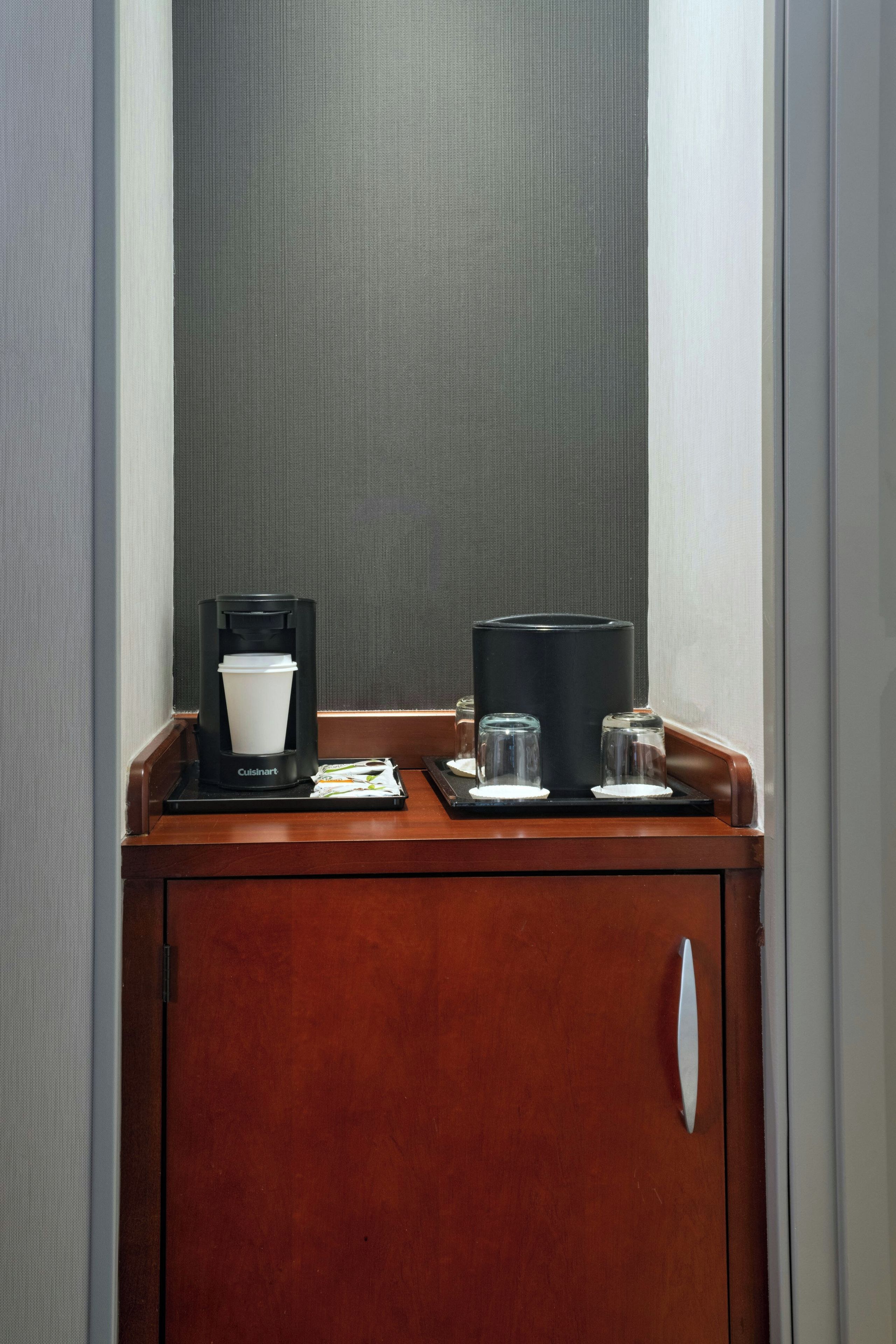 Un mini bar con cafetera, una jarra de vidrio y dos vasos sobre una bandeja de madera.