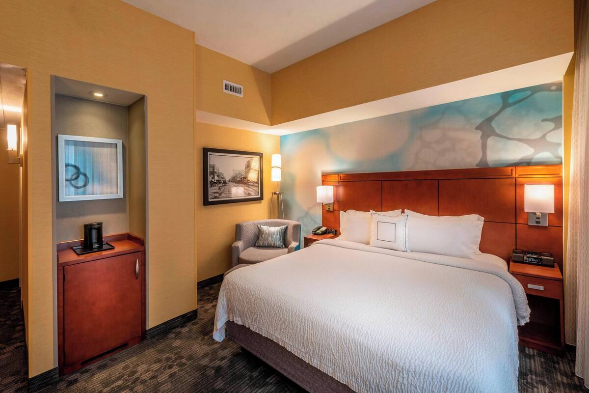 A hotel room with a large bed, a desk with a coffee maker, a chair, and a painting on the wall.