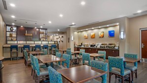 Uma sala de jantar de hotel com várias mesas e cadeiras, balcão de cozinha com alimentos e obras de arte nas paredes.