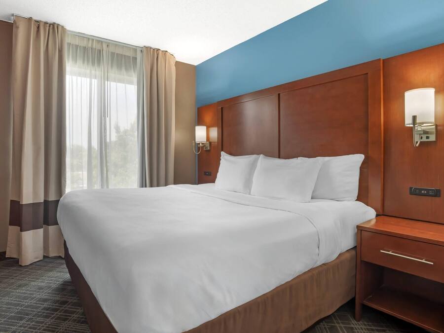 Quarto de hotel com uma cama grande, mesa de cabeceira, luminária na parede e janela com cortinas.