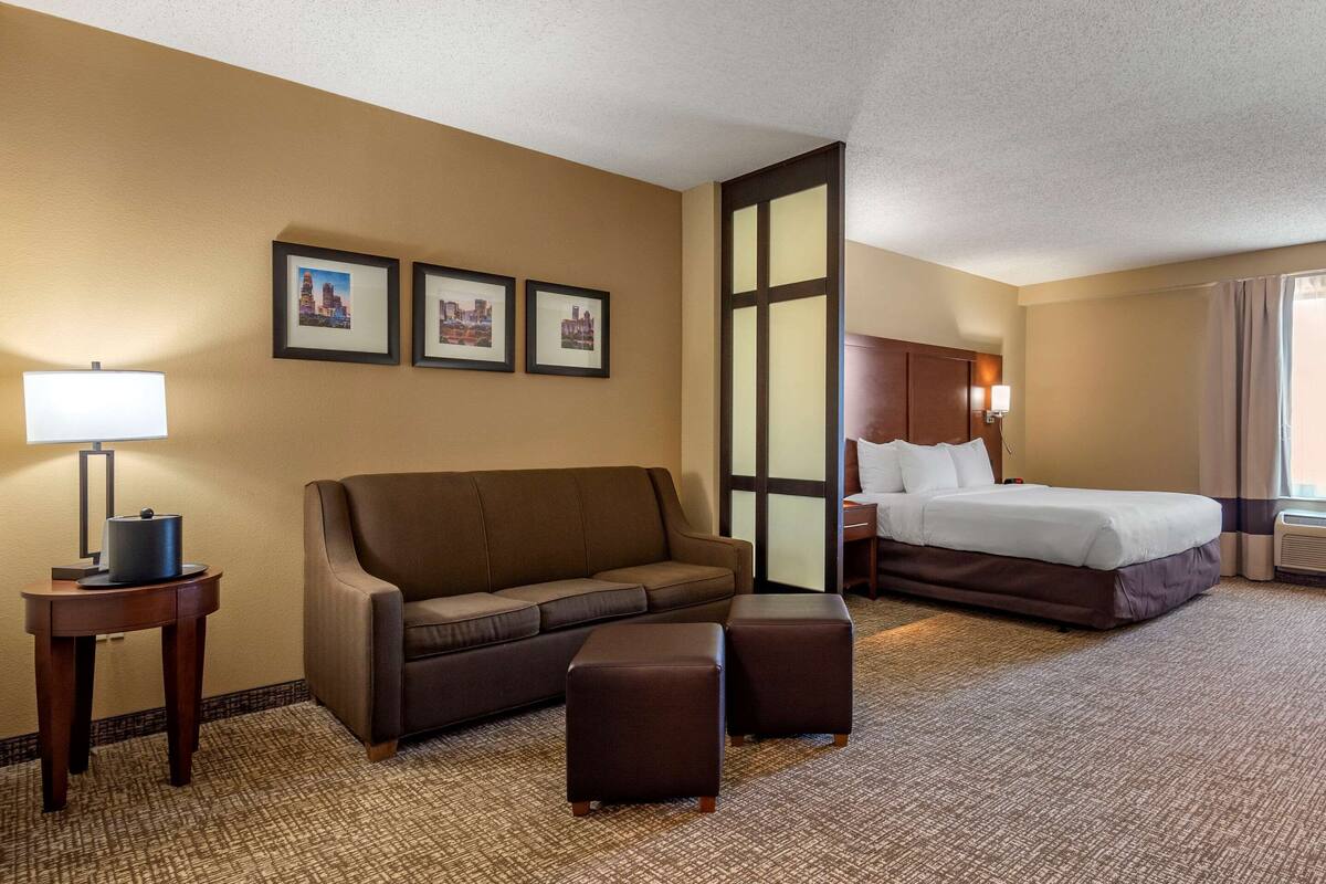 Une chambre d’hôtel avec un canapé marron, un lit, une table de chevet avec une lampe et des tableaux encadrés au mur.