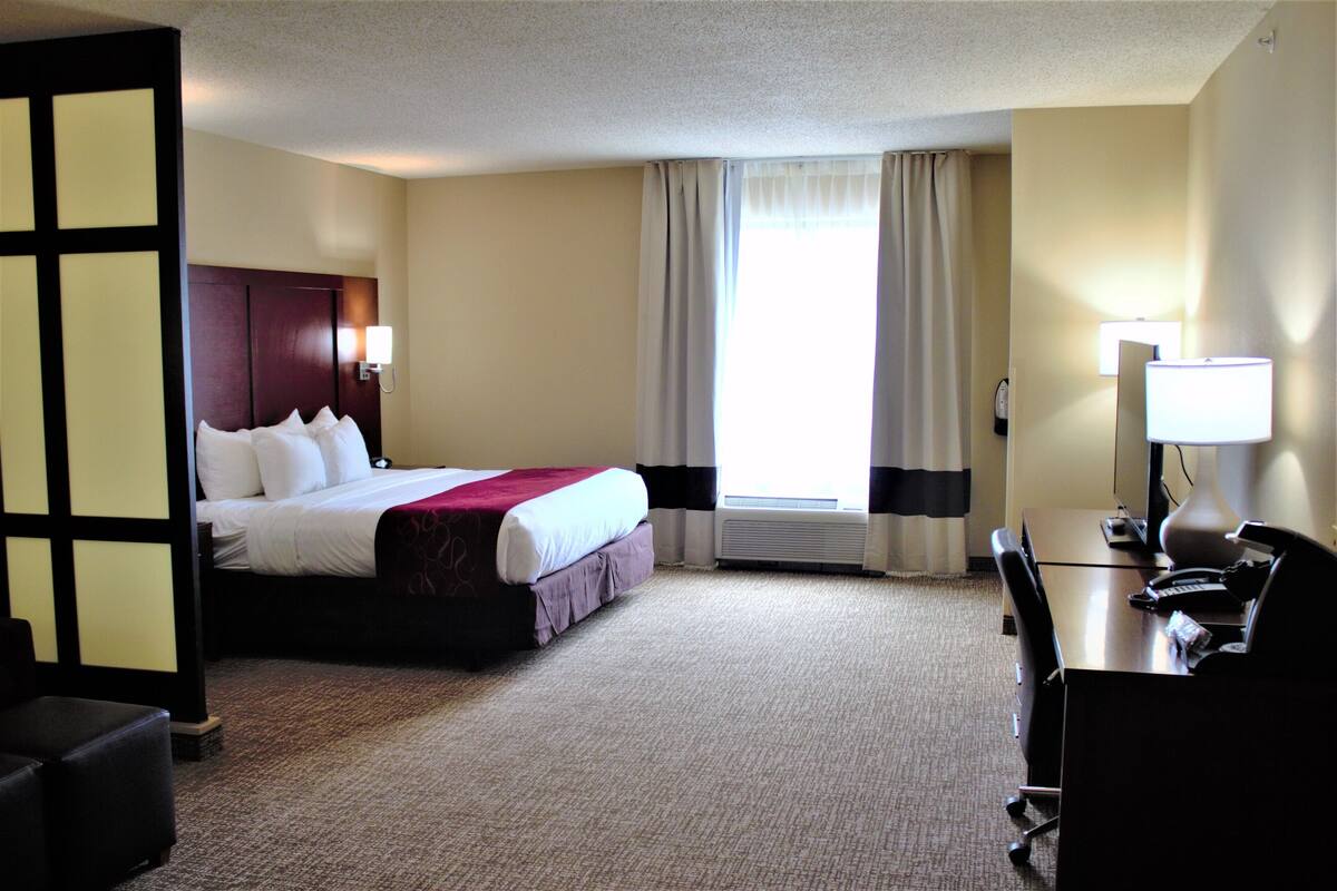 Une chambre d’hôtel avec un lit, un bureau et une chaise, une fenêtre avec des rideaux et une lampe fixée au mur.